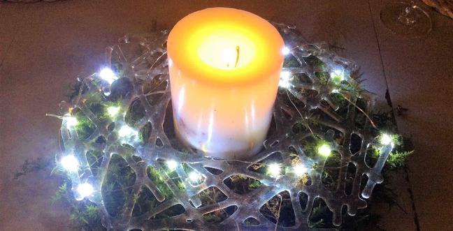 A glass wreath with fairy lights wrapped around it sits on a table with a candle sitting in the middle of it