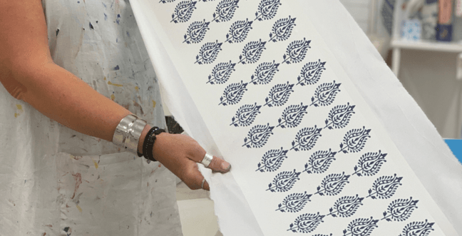 A woman wearing a messy apron and silver rings and bangles holds a long strip of fabric printed with navy blue patterns