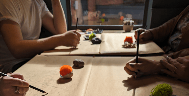 Lit by sunlight through the window next to them four people sit at a table drawing