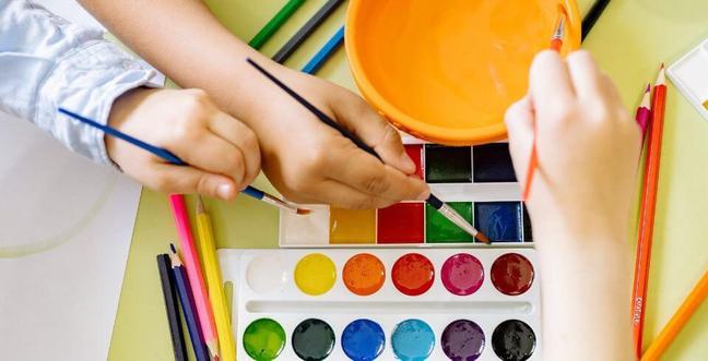 Children's hands over a paint palette with paint brushes and an orange bowl