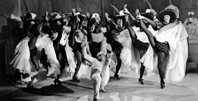 Several female Can-can dancers with one leg in the air surround a man dancing in the middle of them.