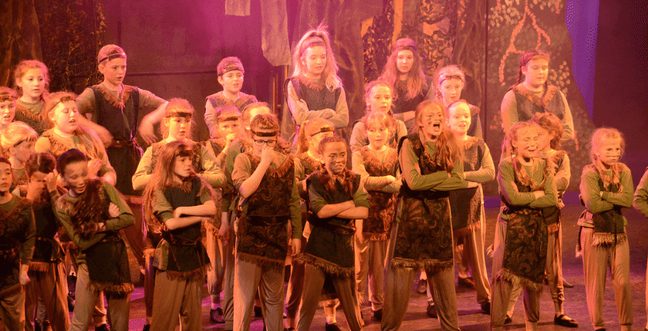 A group of children on stage dressed in green clothes and makeup to appear dirty, with a forest background behind them.