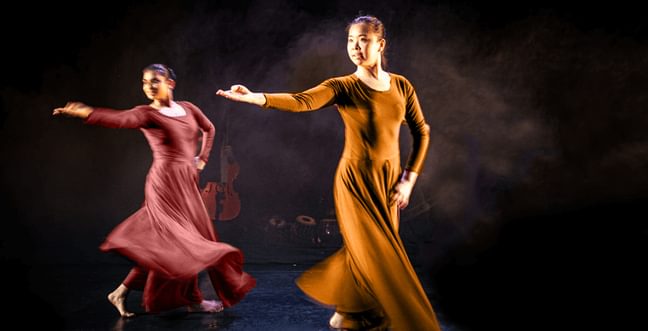 Two female dancers on stage, one in an orange long dress and one in a pink long dress.