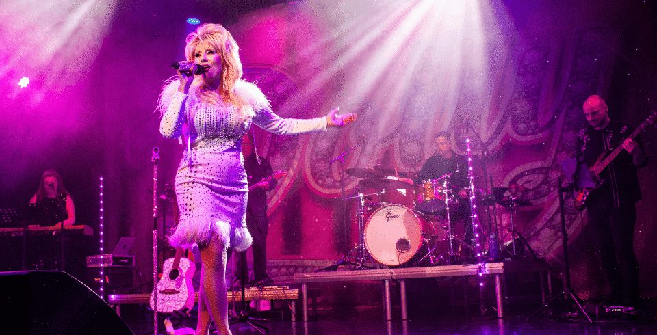 A woman singing on stage wearing a pink dress with frills and feathers, and also a live band behind her
