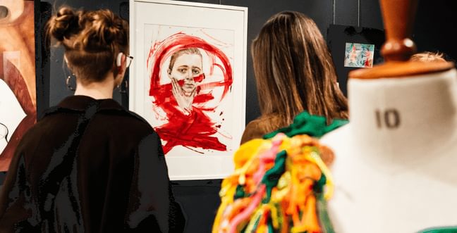 Two women looking at a painting of a person with red streaks of paint over it.